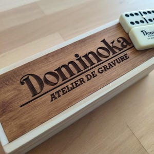 Può includere: Una scatola rettangolare in legno con bordo bianco e un pannello superiore in legno scuro inciso con il testo Dominoka ATELIER DE GRAVURE. Una tessera del domino color bianco sporco con punti neri e il testo Dominoka Atelier de gravure sur dominos et objets.