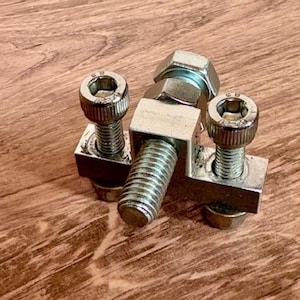 May include: Close-up of several silver metal bolts and nuts. The bolts have hexagonal heads and are attached to square metal plates. The image is taken on a wooden surface.