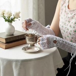 Peut inclure: Gants blancs sans doigts en dentelle avec des liens en ruban. Les gants sont port&eacute;s en tenant une tasse et une soucoupe en argent. Un vase blanc avec des roses blanches est pos&eacute; sur une table recouverte d'une nappe blanche.