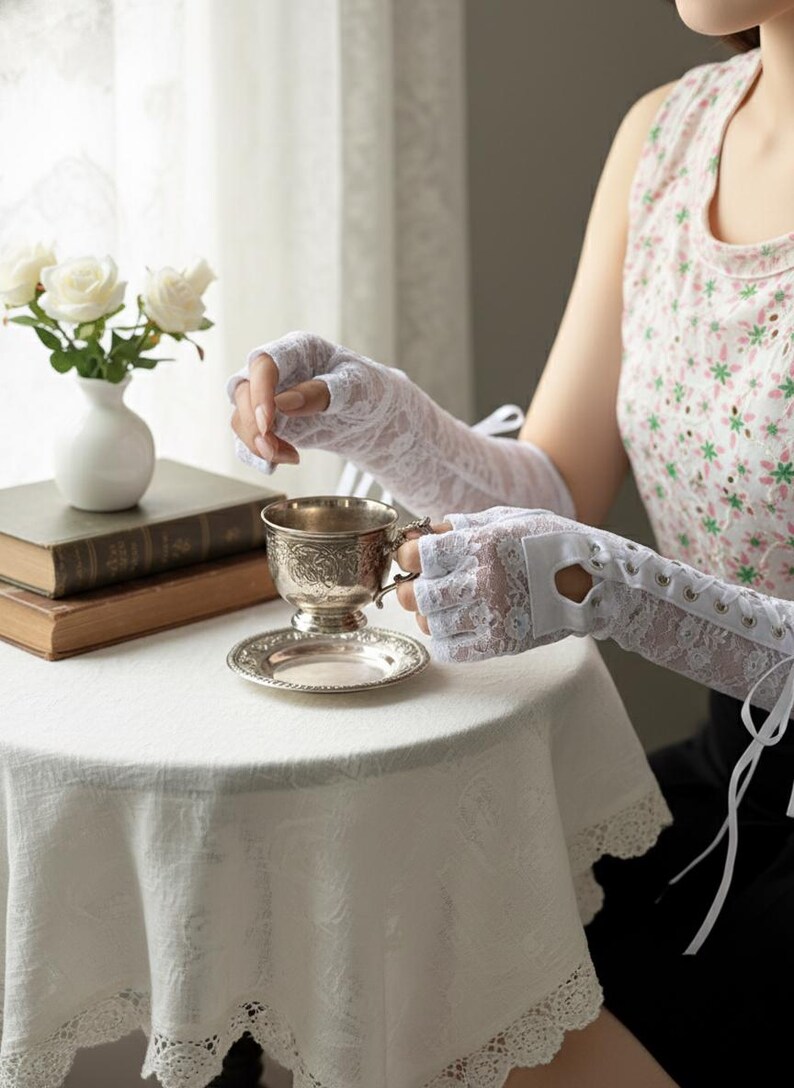 Peut inclure: Gants blancs sans doigts en dentelle avec des liens en ruban. Les gants sont port&eacute;s en tenant une tasse et une soucoupe en argent. Un vase blanc avec des roses blanches est pos&eacute; sur une table recouverte d'une nappe blanche.