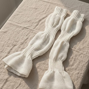 May include: Two off-white, knitted arm warmers with a flared, ruffled cuff. The arm warmers are made of a textured knit fabric and are lying on a beige, textured surface. The arm warmers have a ribbed design at the top and along the arm.