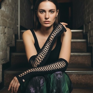 May include: A woman wearing a black tank top and long black arm warmers with a cut-out design. She is sitting on a staircase, wearing a green and purple patterned skirt. The background is a dimly lit brick wall.