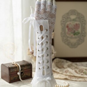 Peut inclure: Gants blancs en dentelle avec un design de style corset sur l'avant-bras. Les gants pr&eacute;sentent un motif de dentelle florale d&eacute;licate et sont orn&eacute;s de rubans de satin et d'une bordure en dentelle &agrave; volants. Les gants sont expos&eacute;s sur une main de mannequin.