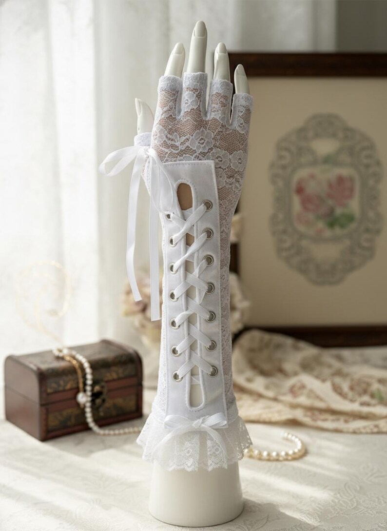 Peut inclure: Gants blancs en dentelle avec un design de style corset sur l'avant-bras. Les gants pr&eacute;sentent un motif de dentelle florale d&eacute;licate et sont orn&eacute;s de rubans de satin et d'une bordure en dentelle &agrave; volants. Les gants sont expos&eacute;s sur une main de mannequin.
