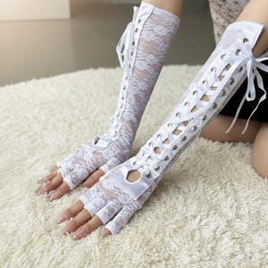Peut inclure: Gants blancs sans doigts en dentelle avec un motif floral et des liens en ruban de satin. Les gants s'&eacute;tendent jusqu'au coude et comportent des &oelig;illets pour le ruban. Les gants sont en dentelle d&eacute;licate.
