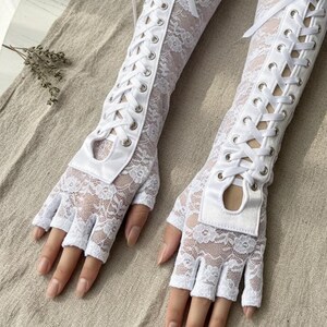 Peut inclure: Une paire de gants blancs sans doigts en dentelle avec un design de style corset. Les gants pr&eacute;sentent un motif floral en dentelle et sont lac&eacute;s sur le bras avec un ruban blanc. Les gants ont une petite ouverture &agrave; la base des doigts.