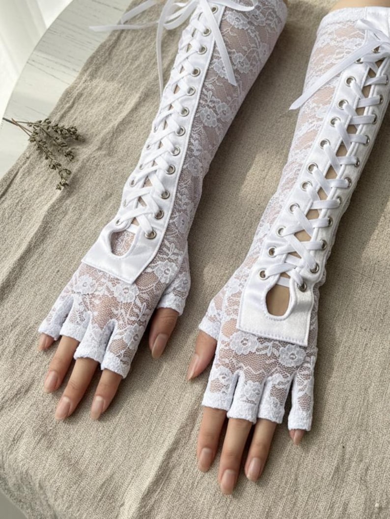 Peut inclure: Une paire de gants blancs sans doigts en dentelle avec un design de style corset. Les gants pr&eacute;sentent un motif floral en dentelle et sont lac&eacute;s sur le bras avec un ruban blanc. Les gants ont une petite ouverture &agrave; la base des doigts.