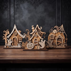 Op de afbeelding: Drie gelaagde houten tafeldecoraties met Halloween-thema's. De set bevat twee huizen en een pompoenstructuur, elk versierd met uitgesneden figuren van geesten, spinnenwebben, uilen en pompoenen, vervaardigd uit lichtgekleurd hout.