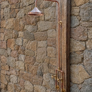 May include: A copper shower fixture mounted on a vertical wooden beam. The unit features a wide, conical shower head, a long vertical pipe, and two brass cross-handle valves at the base, all connected by copper piping and brass fittings.