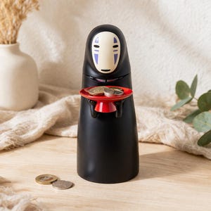 Peut inclure: Une tirelire noire en forme de personnage de Spirited Away, tenant un plateau rouge avec des pièces. La tirelire a un visage blanc avec des marques violettes. Des pièces supplémentaires sont sur la surface en bois.