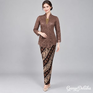 Puede incluir: Una blusa de encaje marrón de manga larga con un broche dorado en el escote, combinada con una falda larga hasta el suelo con un patrón de estilo batik en negro y marrón. El conjunto se presenta con el texto Ganya Delisha en la esquina.