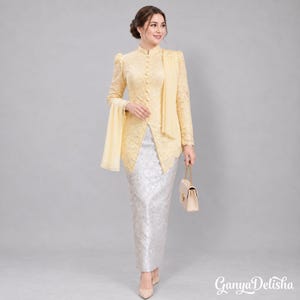 Pode incluir: Um top de manga comprida em renda amarelo-pálido com gola alta, fecho de botões frontal e tecido drapeado a condizer, combinado com uma saia comprida prateada com padrão. O conjunto inclui uma pequena mala bege e tem a etiqueta Ganya Delisha.