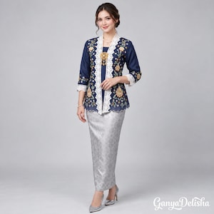 Puede incluir: Una blusa kebaya azul marino de manga larga con bordados florales en dorado y blanco, decorada con encaje blanco en la abertura frontal y los puños. Se combina con una falda larga de tono plateado con un sutil patrón de jacquard.