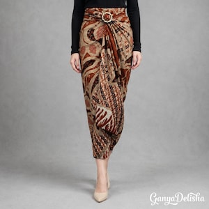 May include: A long wrap skirt featuring a brown and beige batik pattern with intricate geometric and floral motifs. The skirt includes a gold-toned circular buckle at the waist, creating a draped effect with a front fold. The text GanyaDelisha is visible in the bottom right corner.