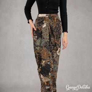 Op de afbeelding: Een lange rok met een donkerbruin en zwart batikpatroon met ingewikkelde bloem- en bladmotieven. De rok heeft een voorpand in wikkelstijl met een gestructureerde tailleband en wordt gecombineerd met een effen zwart shirt met lange mouwen.
