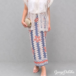 Puede incluir: Una falda larga y recta con un patrón ondulado en azul y blanco, motivos florales naranjas y un borde decorativo naranja en el dobladillo. La falda se combina con un top de encaje blanco y un pequeño bolso de mano redondo dorado.