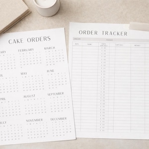 May include: Two white sheets of paper with text, one labeled "Cake Orders" and the other "Order Tracker." The "Cake Orders" sheet displays a calendar layout. A white pen rests on the right side. The background is a neutral, light color.