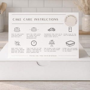 May include: A white cake box with a card on top detailing cake care instructions. The card has icons and text about handling, transporting, and storing a cake. The background features kitchen decor.