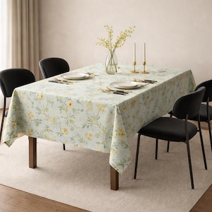 Pode incluir: Uma toalha de mesa retangular com um padrão floral amarelo e verde sobre um fundo azul claro. A mesa está posta com pratos, talheres e um vaso de flores. Quatro cadeiras pretas circundam a mesa.