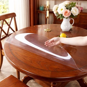May include: A transparent, oval table protector with a white edge is placed on a round, wooden table. The table is a rich, warm brown colour. A hand is shown placing the protector on the table. The table is surrounded by wooden chairs.