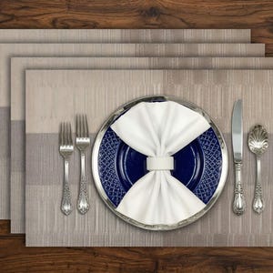 Puede incluir: Un juego de manteles individuales rectangulares de color beige con una sutil textura de rayas horizontales. En el centro, un plato azul oscuro con borde decorativo sostiene una servilleta blanca plegada con un anillo, flanqueado por dos tenedores, un cuchillo y una cuchara de plata.