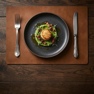 May include: A seared scallop served on a bed of green pea puree with bacon bits and microgreens on a dark gray plate. A silver fork and knife are placed on a brown leather placemat. The setting is arranged for a single dining experience.