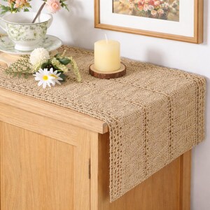 May include: A beige burlap table runner with a woven, openwork pattern. A cream candle rests on a wooden coaster. A teacup and saucer, plus a small floral arrangement, are also visible.