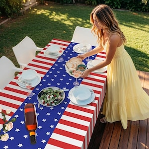 May include: A rectangular tablecloth featuring a patriotic design with red and white horizontal stripes on the sides and a central blue panel adorned with white stars. The table is set with white plates, glassware, a salad bowl, and bread.
