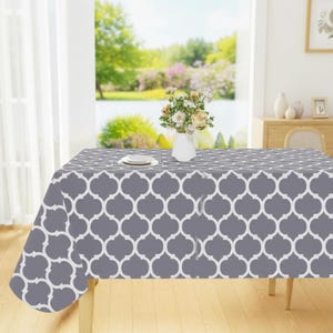 May include: A gray tablecloth with a white quatrefoil pattern covers a wooden table. A white vase with flowers and a plate are on the table. The background shows a window with a view of a lake and trees.