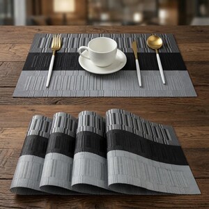 Puede incluir: Un juego de manteles individuales rectangulares con un patrón de rayas en tonos negro, gris oscuro y gris claro. Los manteles tienen una apariencia tejida y texturizada. Uno está extendido con una taza y plato blancos, un tenedor y una cuchara dorados.