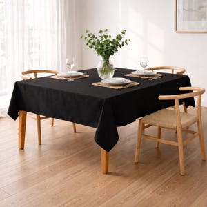 Peut inclure: Une nappe carrée noire recouvre une table en bois dressée pour quatre personnes. La table est entourée de quatre chaises en bois. Chaque couvert comprend une assiette blanche, un verre et un dessous de plat tressé. Un vase de verdure est au centre.