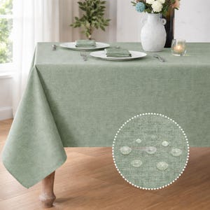 May include: A sage green tablecloth covers a wooden table, set with white plates, napkins, and silverware. A vase of flowers and a lit candle add to the dining setting. The fabric has a textured appearance.