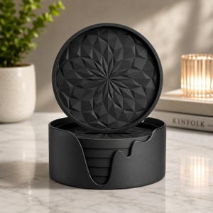 May include: A set of circular black coasters featuring a geometric, faceted starburst pattern on the surface. The coasters are stacked inside a matching black holder with a wavy, open-front design, providing a stable base for the collection.