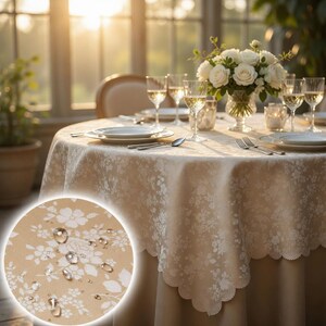 Könnte beinhalten: Ein runder beigefarbener Tischläufer mit weißem Blumenmuster, gedeckt für ein formelles Abendessen. Der Tisch ist mit Tellern, Besteck und Weingläsern gedeckt. Eine Vase mit weißen Rosen steht in der Mitte.