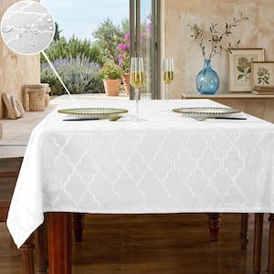 May include: A white tablecloth with a subtle, repeating quatrefoil pattern covers a wooden table. The tablecloth is set with gold-rimmed plates, silverware, and champagne flutes. A blue vase with greenery and framed botanical prints are in the background.