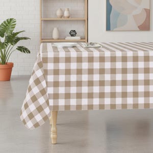 May include: A tan and white checkered tablecloth covers a wooden table. The tablecloth has a classic gingham pattern. A potted plant and decorative items are visible in the background. The table is set with a plate and silverware.