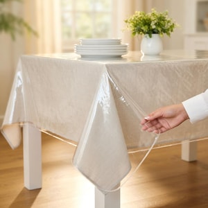 Può includere: Una tovaglia trasparente rettangolare sopra una tovaglia di lino beige su un tavolo bianco. Una pila di piatti bianchi e un vaso bianco con piante verdi sono sul tavolo. Una mano solleva il bordo della tovaglia trasparente.