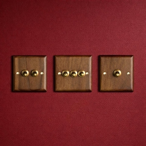 Könnte beinhalten: Drei Lichtschalterplatten aus Holz mit Messing-Kippschaltern und kleinen Messingschrauben. Die Platten sind dunkelbraun und an einer roten Wand montiert. Die Schalter sind in verschiedenen Konfigurationen angeordnet.
