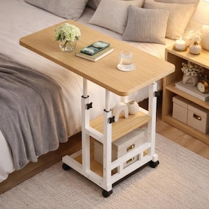 May include: A light wood-tone adjustable bedside table with a white frame and two shelves. The table has a rectangular top with rounded corners, and sits on four wheels. A glass of water, a phone, and a book are on the table.