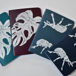 May include: Four notebooks with unique designs. Two notebooks feature white leaf prints on teal and burgundy covers. The other two notebooks have white insect illustrations on navy and teal covers. The notebooks are arranged on a white surface.