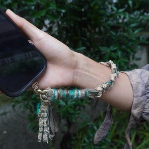 May include: A wrist strap for a mobile phone made of braided and wrapped fabric in shades of green, beige, and brown. The strap features a metallic gold-toned lobster clasp attachment and frayed fabric ends, providing a secure way to hold a device.