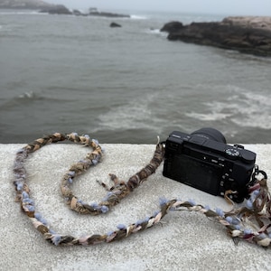May include: A black digital camera attached to a long, decorative camera strap. The strap is crafted from braided fibers in shades of brown, tan, and light blue, featuring a textured, multi-strand design that loops across the surface.