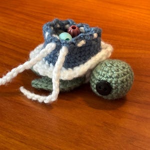 May include: A handmade crochet turtle with a blue and white shell, green body, and a black button eye. The turtle's shell is open, revealing colorful beads inside. White yarn is used for the turtle's head and legs.