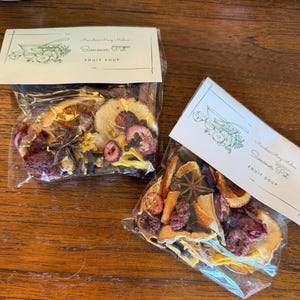 May include: Two clear plastic pouches containing dried fruit soup ingredients, including orange slices, star anise, cloves, and berries. Each bag features a white paper header with a line drawing of a bowl and the text Simmer Pot FRUIT SOUP.