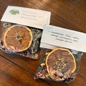 May include: Two clear plastic pouches containing dried orange slices, pine, cedar, eucalyptus, sage, and juniper berries. The labels read Marvelous Mary Malone Simmer Pot Sweater Weather and list ingredients including pine, cedar, orange, eucalyptus, sage, and juniper berries.