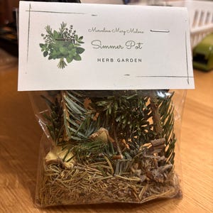 May include: A clear plastic bag filled with dried herbs and pine needles, topped with a white label. The label reads "Marvelous Mary Malone Simmer Pot HERB GARDEN" with a green illustration of herbs.