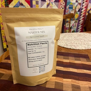 May include: A brown paper bag of "Master Mix" sprouted spelt flour. The label includes nutritional information and the text "Marvelous Mary's". The bag is on a wooden cutting board with a checkered pattern.