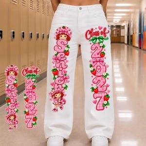 May include: White denim jeans featuring a vertical graphic print on each leg. The left leg displays the word Senior with a cartoon character and strawberries. The right leg shows the text Class of 2027 with strawberries and pink hearts.