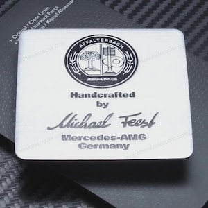 Puede incluir: Una placa metálica rectangular de tono plateado que presenta el logotipo negro de Affalterbach AMG en la parte superior. Debajo del logo, el texto dice Handcrafted by Michael Feest, seguido de Mercedes-AMG Germany en una fuente sans-serif.