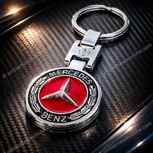 Puede incluir: Un llavero plateado de Mercedes-Benz con un emblema rojo y plateado. El emblema presenta el logotipo de Mercedes-Benz, una estrella de tres puntas y las palabras "Mercedes" y "Benz". El llavero tiene un anillo y un cierre plateados.